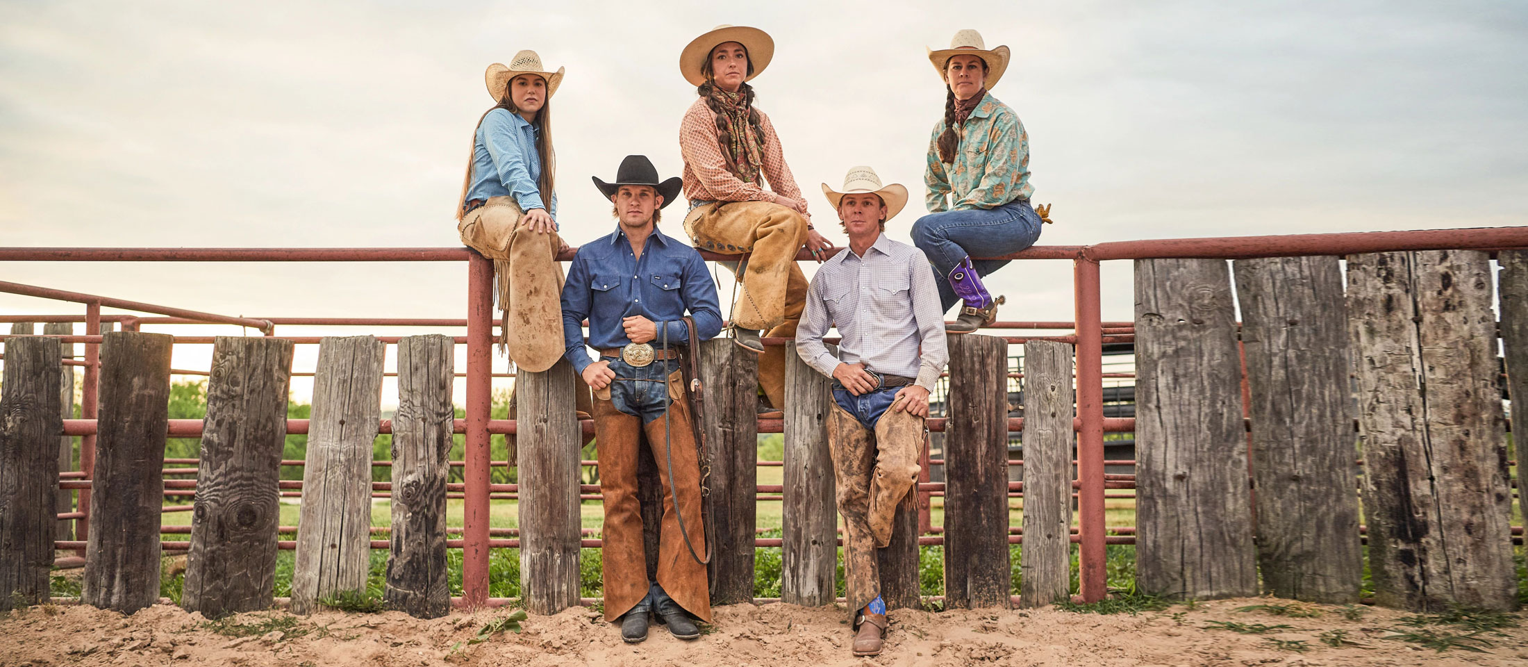 Tres mujeres y dos hombres en un rancho vestidos con jeans, camisa, sombrero de vaquero y botas de vaquero.